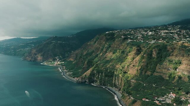 A smooth aerial drone shot capturing the southern coastline of Madeira Island, filmed near Cascata dos Anjos. The camera faces toward the western part of the island, revealing rugged volcanic cliffs
