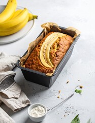 A freshly baked banana bread in a loaf pan, topped with banana slices, placed on a clean kitchen surface. A bunch of ripe bananas and a small bowl of flour are visible, adding to the warm, homey feel.
