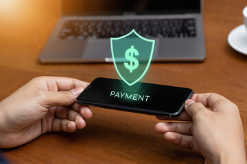 Hands holding smartphone with payment screen and green dollar sign shield above it on wooden table with laptop in background