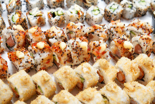 Close-up of assorted sushi rolls arranged in rows, topped with sesame seeds, spicy sauce, and mayonnaise. A variety of maki and uramaki sushi with fresh ingredients such as salmon, cucumber, and rice.