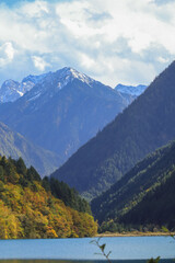 Fototapeta premium Serene mountain landscape with tranquil lake in foreground, surrounded by lush forests and majestic snow capped peaks under partly cloudy sky