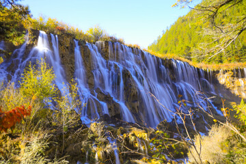 Stunning waterfall cascades over rocky cliffs surrounded by lush greenery and vibrant autumn foliage, creating serene and picturesque natural landscape © pirom