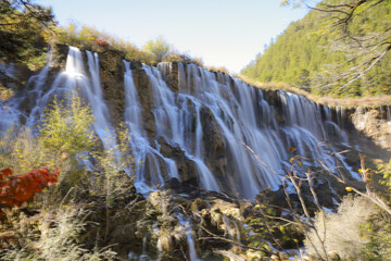 Majestic waterfall cascades over rugged rocks surrounded by lush greenery and autumn foliage, creating serene and picturesque natural scene © pirom