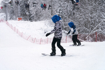 A picturesque ski slope with two teenage snowboarders against the backdrop of a chairlift and frost-covered trees.