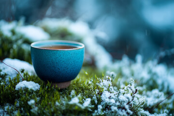 teal cup filled with tea resting on a moss-covered surface with snow