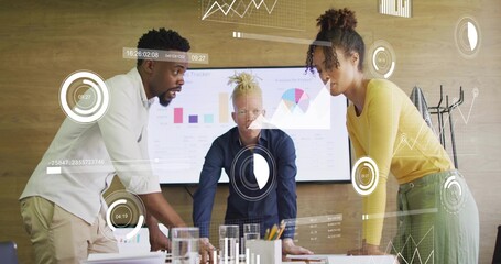 Leaning three colleagues in suits reviewing charts at table with screen display, HUD overlays