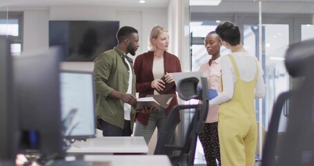 Reviewing documents, four coworkers in bright clothes checking laptop tablet papers in open office