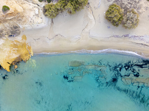 Aerial view of the turquoise sea gently kisses the sandy shore, embraced by rugged cliffs and scattered trees, Paralia Mitikas, Milos, Greece.
