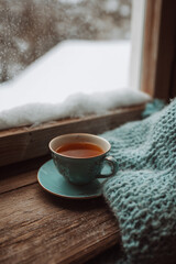 winter composition tea, cup of tea resting on a wooden surface with a soft blanket nearby, viewed through a snowy window