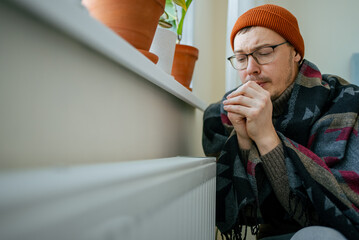 Man wearing warm clothes and a blanket, rubbing hands together for warmth, sitting by a cold radiator inside his home, facing increased energy bills and winter struggles