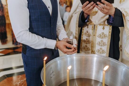 Cropped shot of a man in a vest holding holy oil near a baptismal font with lit candles, beside a priest reading a holy book during a Christian baptism ceremony