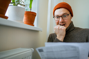 Stressed man in warm hat looks shocked, biting nails while reading an unexpectedly high heating bill, overwhelmed by rising living costs, debt and energy crisis