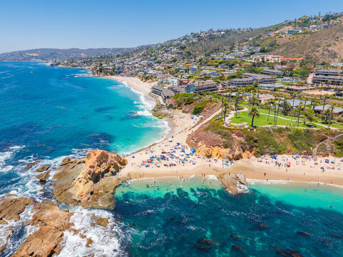 Aerial view of the coastline where white sands meet the turquoise sea, dotted with beachgoers and framed by lush greenery, Laguna Beach, California, United States.