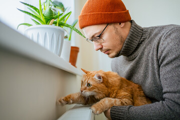 Man and ginger cat sharing warmth by a radiator, both experiencing comfort and connection, representing companionship and saving money on heating during cold days