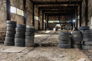 Multiple stacks of used, dirty car tires inside a large, abandoned industrial warehouse with concrete walls and strong shadows © AndreyZayats