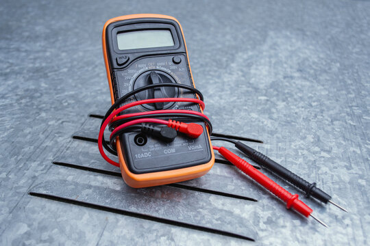 Orange digital multimeter with red test probe leads on textured gray metal background. Professional electrical measuring instrument for voltage and electrical work.