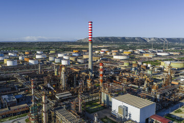 Aerial view of a sprawling industrial complex with a towering red and white chimney piercing the skyline amidst a network of pipes and tanks, Syracuse, Sicilia, Italy.