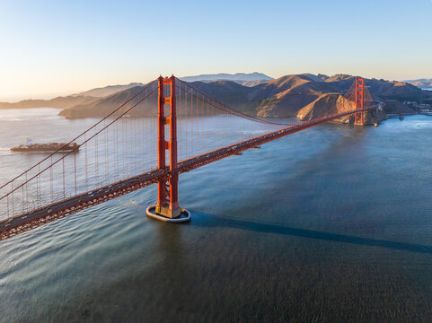 Aerial view of the iconic red Golden Gate Bridge stretching across the bay, with sunlight glinting on the water and hills in the background, San Francisco, California, United States.