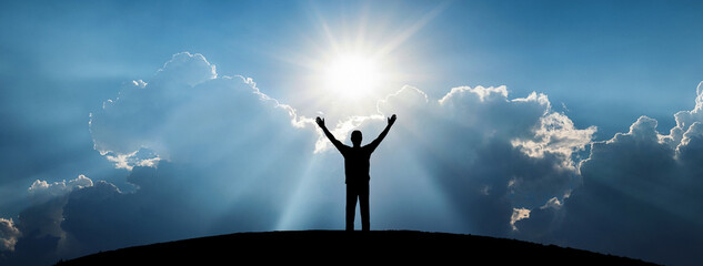 Human silhouette standing on a hill with raised arms against bright sun rays breaking through...