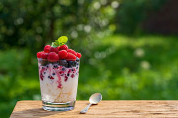 Summer dessert for breakfast in a glass of cottage cheese with sour cream, blueberries, raspberry, jam and honey on nature background, copy space. Healthy diet with fresh berries and dairy product