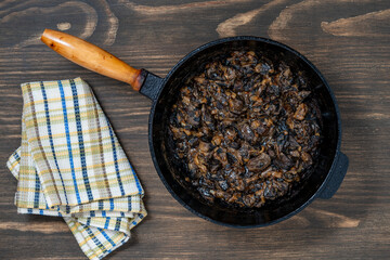 Cast iron skillet with fried wild mushrooms with onions and sour cream on wooden background, top view, copy space, closeup