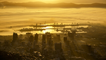 View of the city skyline and harbor shrouded in a golden mist, the sun's rays piercing through the clouds, casting long shadows, Cape Town, South Africa.
