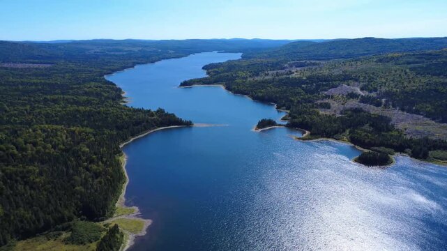 Breathtaking aerial view of a winding lake flowing through lush forests and gentle mountains, reflecting sunlight under a blue sky. Lake Mitis, Quebec, Canada, 2025.