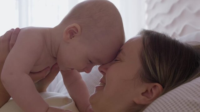 mother lifting baby above chest on bed. morning exercise and play mom with infant with bright window light. cheerful movement and eye contact. happy domestic maternal routine. cozy bonding