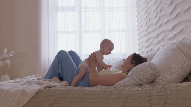 mother lifting baby above chest on bed. morning exercise and play mom with infant with bright window light. cheerful movement and eye contact. happy domestic maternal routine. cozy bonding