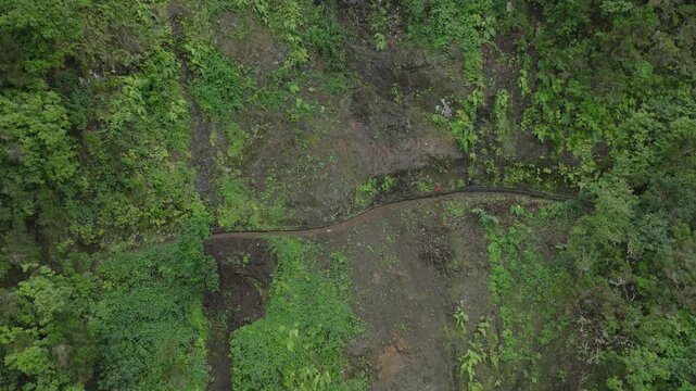 A drone shot of the PR9 Levada trail carved into a steep vertical cliff on Madeira Island. Dense levada vegetation covers the rock wall, while the narrow water channel and walking path are are clearly