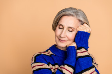 Aged mature woman in a blue knit sweater rests her head on her hands with a peaceful smile against a warm beige background