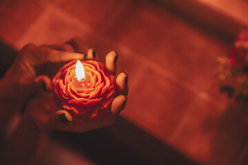 Close up of woman's hands gently holding a burning red rose low-temperature massage candle in warm dim light.