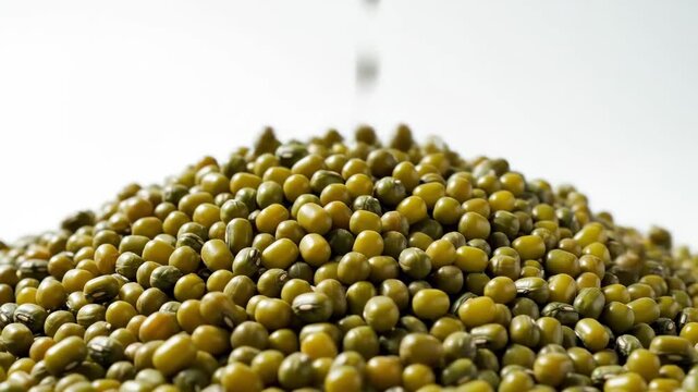 Mung Beans Pouring into Pile - A close-up shot of mung beans falling onto a pile of more mung beans, against a white background. The small, round, green beans are densely packed.