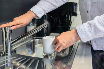 Everyday Utility close view of woman filling mug with water at office sink, running faucet, stainless steel basin and cups visible, basic hydration task in workplace kitchen