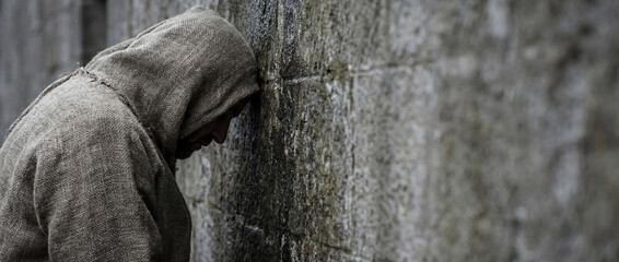 Man wearing sackcloth robe leaning head against ancient stone wall. Biblical concept of repentance, mourning and despair. Dramatic portrait of a prophet or sinner praying.