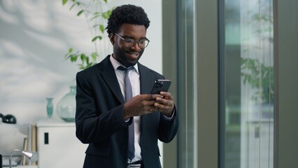 Smiling African American businessman using smartphone in office business man ethnic male guy looking at mobile phone cheerful professional entrepreneur checking messages browsing internet success