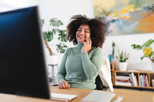 A woman with curly hair smiles while talking on her phone at her desk, with a computer and plants in the background.