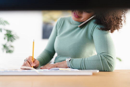 A woman with curly hair talks on her phone while writing in a notebook with a pencil at her desk.