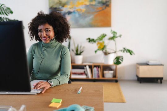 A smiling woman with curly hair wears a headset and works at a computer in a bright, modern office space with plants and artwork.