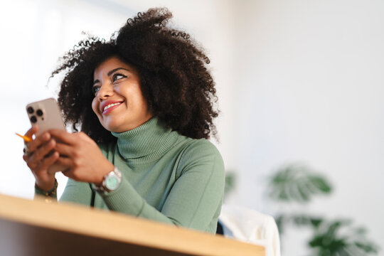 A smiling woman with curly hair holds a smartphone and a pencil, looking away from the camera.