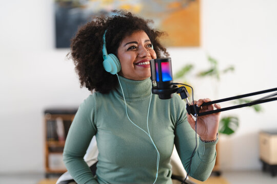A woman with curly hair wears headphones and smiles while speaking into a microphone, recording audio for a podcast or broadcast.