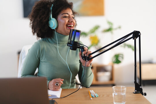 A woman with curly hair wearing headphones speaks into a microphone, recording audio for a podcast or broadcast.