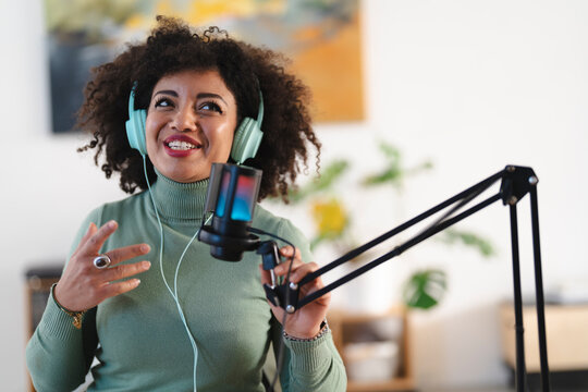 A woman with curly hair wears headphones and speaks into a microphone, gesturing with her hand. She is recording audio content in a studio setting.