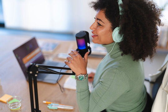 A woman with headphones speaks into a microphone while recording a podcast at her desk with a laptop and notes.