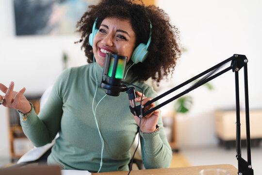 A woman with curly hair wearing headphones speaks into a microphone, gesturing with her hands while recording a podcast or broadcast.