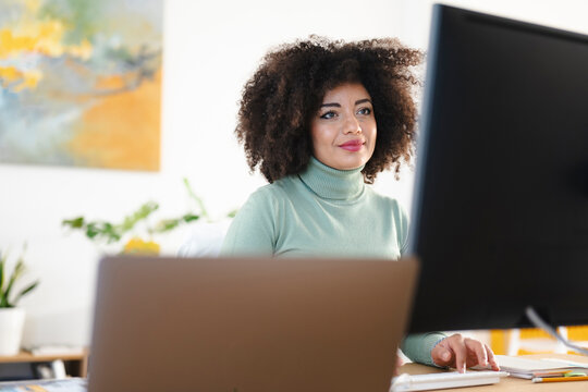 A woman with curly hair works on her computer in a bright office. She is focused on her screen, typing on a keyboard.