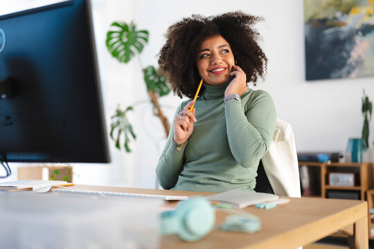 A woman with curly hair smiles while talking on her phone and holding a pencil at her desk.