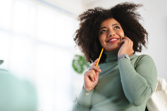 A woman with curly hair smiles while holding a pencil and talking on her phone, looking up thoughtfully.