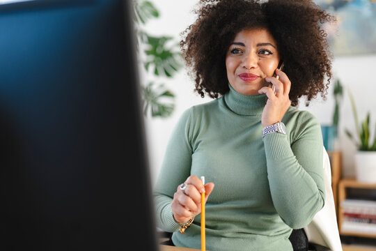 A woman with curly hair talks on her phone while holding a pencil, working at her desk with a computer in front of her.