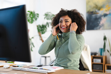 A smiling woman with curly hair adjusts her headset while working at her desk, looking at a computer screen.